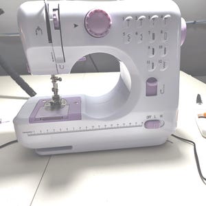 May include: A white and lavender sewing machine with a handle, dials, and stitch selection options. The machine has a built-in ruler and an on/off switch. The machine is designed for basic sewing tasks.