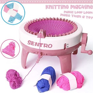 May include: Pink knitting machine with a white base and a crank handle. The machine is making a pink knitted fabric. The image also shows a pink hat, a pink and blue scarf, and three balls of yarn in pink, blue, and white. The text on the machine reads "Knitting Machine Magic Loop Loom More Than a Toy Senttro".