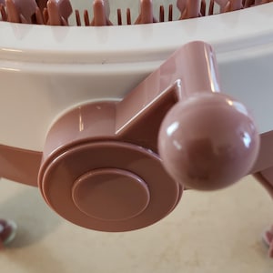 May include: Close-up of a pink plastic knitting loom with a round base and a crank handle. The loom has a white top and a pink bottom. The crank handle is used to turn the loom and knit the yarn.