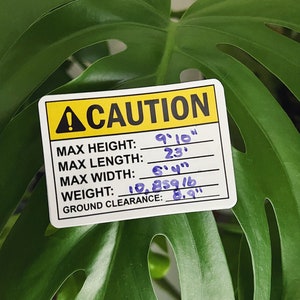 May include: A yellow caution sign with black text and a black exclamation point. The sign lists maximum height, length, width, weight, and ground clearance measurements in inches. The measurements are handwritten in black ink.
