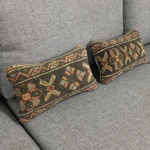 May include: Two rectangular throw pillows featuring a dark brown base with woven geometric patterns in muted shades of green, orange, and cream. The pillows are positioned on a light gray fabric sofa with a visible wooden frame at the top edge.