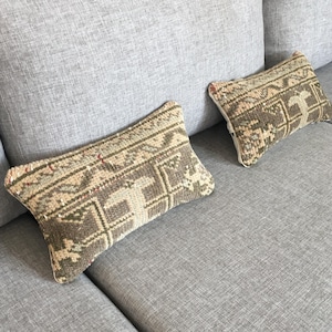 Op de afbeelding: Twee rechthoekige sierkussens met een geweven, veelkleurig geometrisch patroon in tinten bruin, beige en gedempt groen. De kussens hebben een getextureerd, tapijtachtig uiterlijk met een zichtbare pool en liggen op een grijze stoffen bank.