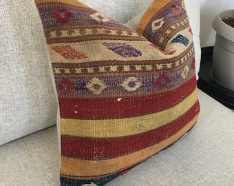 May include: A square throw pillow featuring a woven textile cover with horizontal stripes in shades of red, yellow, orange, and blue, accented by a repeating pattern of small geometric diamond shapes in purple, red, and white across the top section.