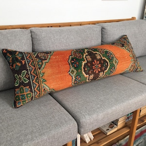 Könnte beinhalten: Ein langes, rechteckiges Kissen mit einem bunten, gemusterten Stoffbezug. Das Kissen ist orange, grün, braun und schwarz mit einem geometrischen Muster. Das Kissen liegt auf einem grauen Sofa.