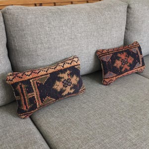 May include: Two rectangular throw pillows with intricate, colorful patterns rest on a gray sofa. The pillows feature a dark blue background with geometric designs in shades of orange, brown, and green. The sofa has a light gray fabric.