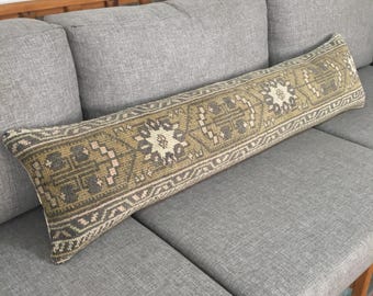 May include: A long, rectangular lumbar pillow featuring a woven pattern in shades of olive green, charcoal gray, and cream. The design consists of repeating geometric motifs and stylized floral shapes arranged in a horizontal row across the surface.
