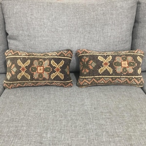 May include: Two rectangular throw pillows featuring a dark charcoal base with intricate woven geometric patterns in muted shades of sage green, dusty rose, and cream. The designs include repeating cross motifs and zigzag borders.