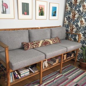 May include: A wooden three-seat sofa with light gray cushions and a long, rectangular bolster pillow featuring a red, cream, and black floral pattern. The sofa frame includes a slatted lower shelf filled with various books and wooden armrests.