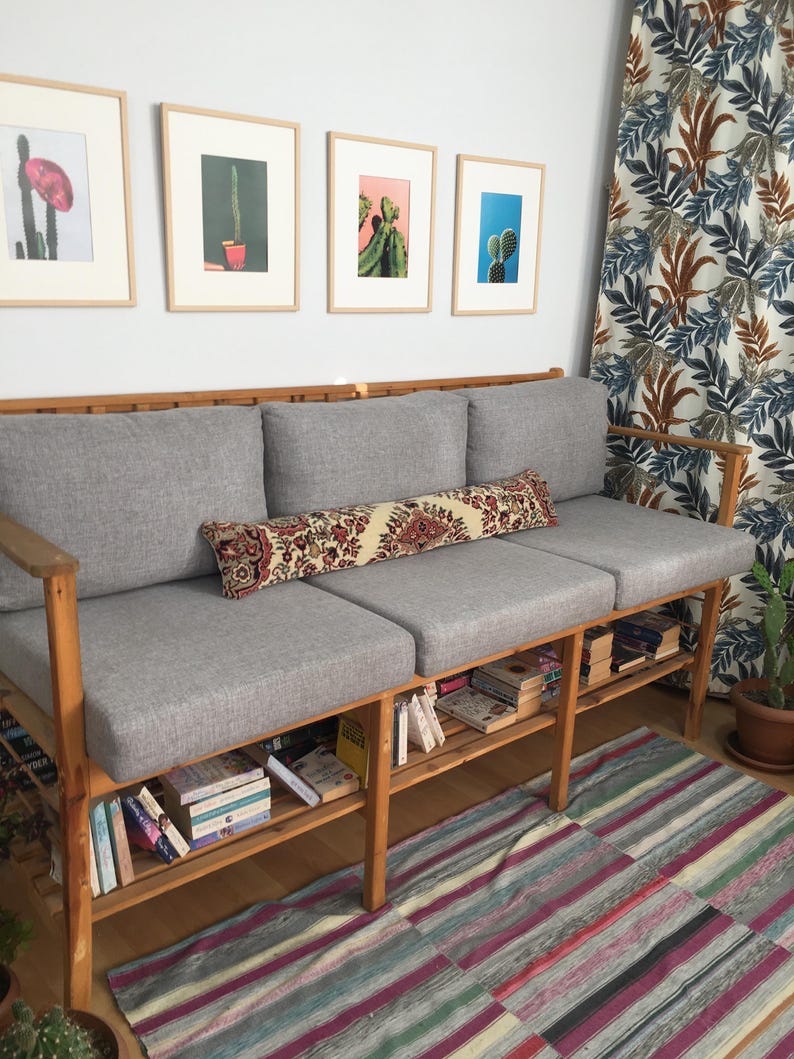 May include: A wooden three-seat sofa with light gray cushions and a long, rectangular bolster pillow featuring a red, cream, and black floral pattern. The sofa frame includes a slatted lower shelf filled with various books and wooden armrests.