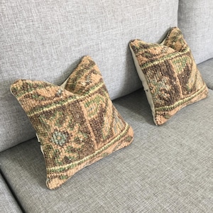 May include: Two square throw pillows featuring a textured, woven surface with a multi-colored pattern in shades of brown, tan, and muted green. The pillows have a thick, knotted pile construction and are displayed side by side on a gray fabric surface.