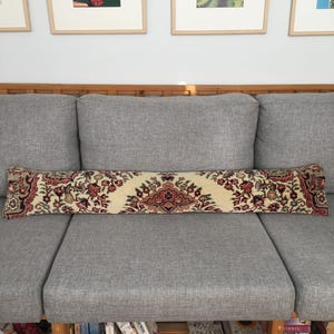 May include: A long, rectangular bolster pillow featuring a cream-colored base with a detailed, symmetrical floral pattern in shades of deep red, dark green, and black, reminiscent of a traditional rug design, positioned horizontally on a gray sofa.