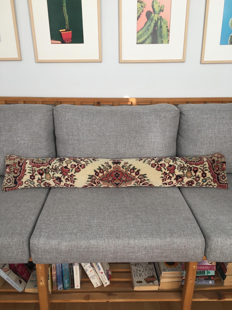 May include: A long, rectangular bolster pillow featuring a cream-colored base with a detailed, symmetrical floral pattern in shades of deep red, dark green, and black, reminiscent of a traditional rug design, positioned horizontally on a gray sofa.