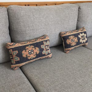 May include: Two patterned throw pillows on a gray couch. The pillows are rectangular and have a dark blue background with intricate patterns in shades of brown, orange, and green.