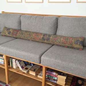 May include: A gray fabric sofa with a long, patterned pillow. The sofa has a wooden frame and is sitting on a wooden floor. There are books on shelves underneath the sofa.