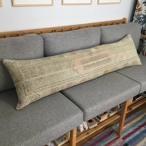 May include: A long, rectangular, off-white throw pillow with a woven pattern and fringe on the ends. The pillow is resting on a grey sofa with wooden legs and shelves.