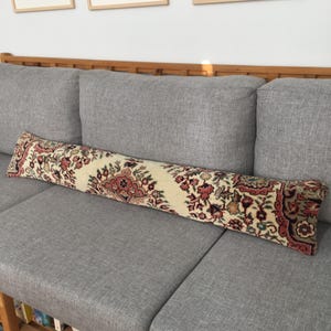 May include: A long, rectangular bolster pillow featuring a cream-colored base with an intricate, repeating floral and geometric pattern in shades of deep red, muted green, and dark brown, reminiscent of a traditional woven rug textile design.
