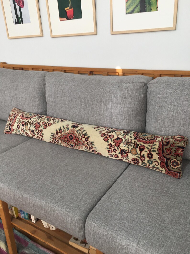 May include: A long, rectangular bolster pillow featuring a cream-colored base with an intricate, repeating floral and geometric pattern in shades of deep red, muted green, and dark brown, reminiscent of a traditional woven rug textile design.