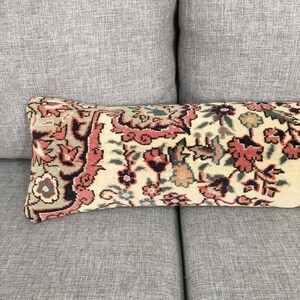 May include: A rectangular lumbar pillow featuring a woven textile cover with a dense floral pattern in shades of muted coral, dark navy blue, and sage green on a cream-colored background, showcasing intricate botanical motifs and ornate borders.