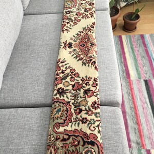 May include: A long, rectangular runner rug featuring a cream-colored background with an intricate floral pattern in shades of dusty rose, sage green, and dark charcoal. The textile displays a repeating symmetrical design with ornate botanical motifs.