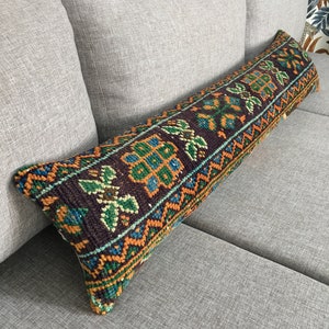 Könnte beinhalten: Ein langes, rechteckiges Kissen mit einem farbenfrohen, geometrischen Muster in Orange-, Grün-, Blau- und Brauntönen. Das Kissen ist aus gewebtem Stoff und hat einen Fransenrand.