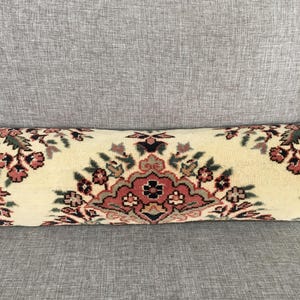 May include: A cylindrical bolster pillow featuring a cream-colored base with a repeating floral pattern in shades of dusty rose, dark green, and black. The design consists of symmetrical botanical motifs arranged horizontally across the fabric.
