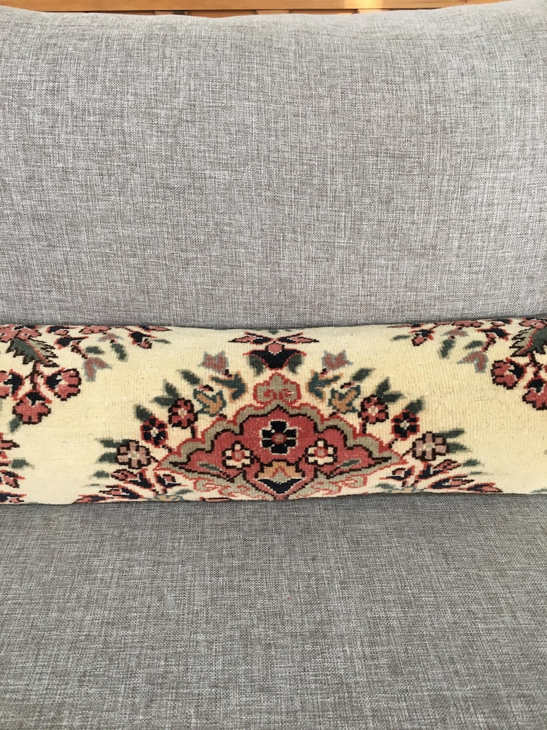 May include: A cylindrical bolster pillow featuring a cream-colored base with a repeating floral pattern in shades of dusty rose, dark green, and black. The design consists of symmetrical botanical motifs arranged horizontally across the fabric.