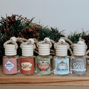 May include: Five small glass bottles with wooden tops and labels featuring text like "Sookie's Christmas Bakes" and "I Smell Snow." The bottles are arranged on a wooden surface, with a festive green garland in the background.