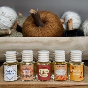May include: Five small glass bottles with wooden lids and labels. The labels read "Sookie's Autumnal Bakes", "Stars Hollow", "Luke's Cinnamon Chai Latte", "Autumn Dragonfly Inn", and "I Am An Autumn". A decorative pumpkin and yarn balls are in the background.