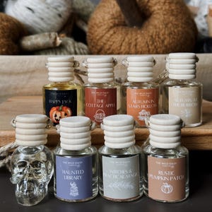 May include: A collection of small glass bottles with wooden lids, featuring various labels with autumn and Halloween themes. The labels include text like "Happy Jack" and "Haunted Library", with pumpkin and skull-shaped bottles. A decorative pumpkin is in the background.