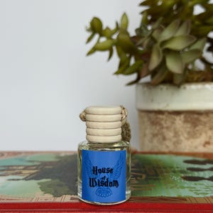 Pu&ograve; includere: Una piccola bottiglia di vetro con tappo in legno ed etichetta blu con la scritta "House of Wisdom" con un disegno alato. La bottiglia &egrave; appoggiata su un libro rosso con scritte dorate, con una pianta in vaso sullo sfondo.