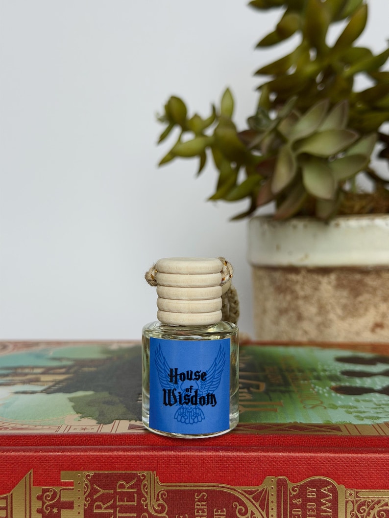 Pu&ograve; includere: Una piccola bottiglia di vetro con tappo in legno ed etichetta blu con la scritta "House of Wisdom" con un disegno alato. La bottiglia &egrave; appoggiata su un libro rosso con scritte dorate, con una pianta in vaso sullo sfondo.