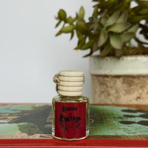 Pu&ograve; includere: Una piccola bottiglia di vetro con tappo in legno ed etichetta rossa con la scritta "House of Courage" e l'illustrazione di un leone. La bottiglia &egrave; riempita con un liquido trasparente e poggia su un libro rosso.