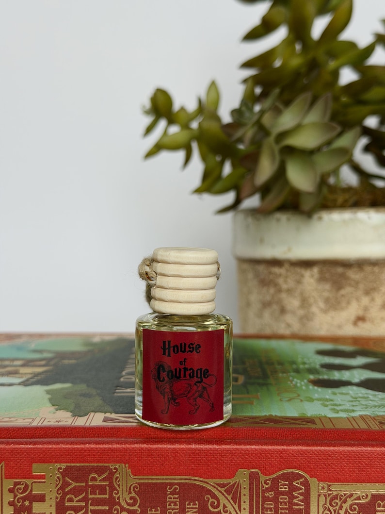 Pu&ograve; includere: Una piccola bottiglia di vetro con tappo in legno ed etichetta rossa con la scritta "House of Courage" e l'illustrazione di un leone. La bottiglia &egrave; riempita con un liquido trasparente e poggia su un libro rosso.