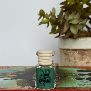 Pu&ograve; includere: Una piccola bottiglia di vetro con un tappo in legno chiaro e un'etichetta verde con la scritta "House of Cunning" e un'illustrazione di un serpente. La bottiglia &egrave; appoggiata su un libro rosso con scritte dorate, con una pianta succulenta sullo sfondo.