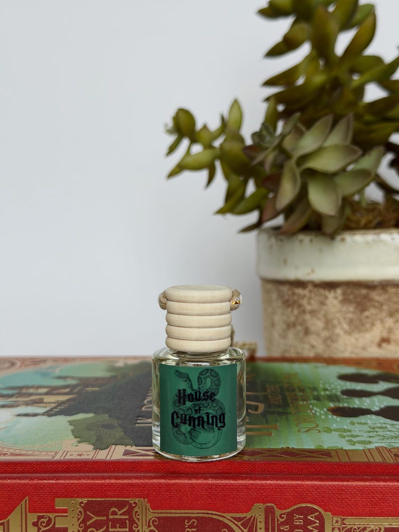 Pu&ograve; includere: Una piccola bottiglia di vetro con un tappo in legno chiaro e un'etichetta verde con la scritta "House of Cunning" e un'illustrazione di un serpente. La bottiglia &egrave; appoggiata su un libro rosso con scritte dorate, con una pianta succulenta sullo sfondo.