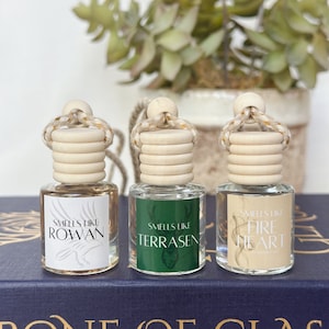 May include: Three small glass bottles with wooden tops and rope hangers. Each bottle has a label with text: "Smells Like Rowan", "Smells Like Terrasen", and "Smells Like Fire Heart". The bottles sit on a blue book with gold lettering.