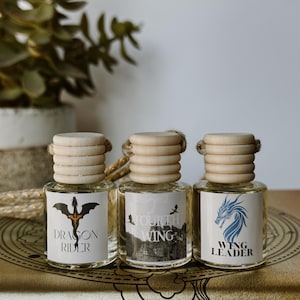 May include: Three small glass bottles with wooden caps and rope accents. Each bottle has a label with a fantasy-themed design and text: "DRAGON RIDER", "FOURTH WING", and "WING LEADER". The bottles are displayed on a gold-colored surface.