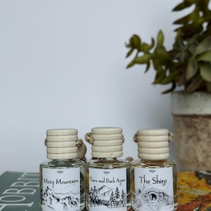 May include: Three small glass bottles with wooden tops and labels featuring black and white illustrations and text. The labels read "Misty Mountains", "There and Back Again", and "The Shire". The bottles are displayed on a book.