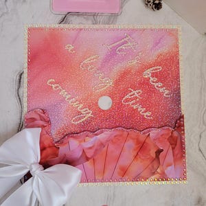 May include: A pink and red decorative item with the text "Coming a long time" in cursive. The item has a white bow and a pink oval with the name "damira" on it. The item is decorated with glitter and rhinestones.