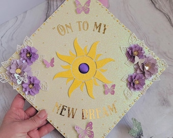 May include: A decorated graduation cap with the text "ON TO MY NEW DREAM". The cap is adorned with a yellow sun, purple flowers, and butterflies. The edges are lined with small pearls.