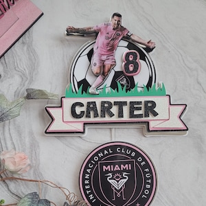 May include: A pink and black cake topper featuring a footballer in a pink jersey with the number 8 on the back. The player is standing on a football with green grass around it. The topper also includes a banner with the name "CARTER" and a pink and black circle with the text "INTERNACIONAL CLUB DE FUTBOL MIAMI MMXX".
