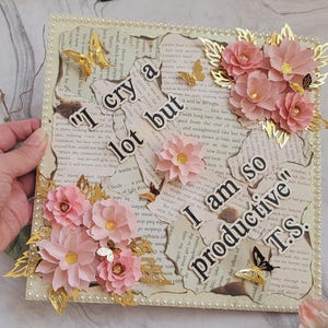 Könnte beinhalten: Dekoratives gerahmtes Kunstwerk mit dem Text "I cry a lot but I am so productive T.S." auf gealtertem Papier. Es zeigt rosa Papierblumen, goldene Schmetterlinge und einen Perlenrand.