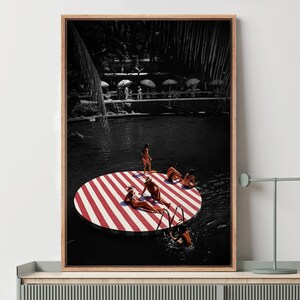 May include: A framed art print depicts a black and white scene with a red and white striped circular platform in a dark body of water. Several people are sunbathing on the platform. The print is displayed on a gray cabinet with a lamp and decorative items.