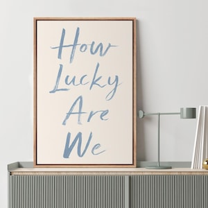 May include: A framed canvas art print with the text "How Lucky Are We" in blue script. The artwork is displayed on a light-colored wall above a gray cabinet with vertical lines. A modern lamp and a gold sphere are on the cabinet.