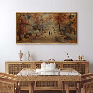 May include: A framed autumn-themed painting depicts a street lined with houses and trees in fall colors. Pumpkins are scattered along the road. Figures of people are walking on the street. The painting is in a gold frame.