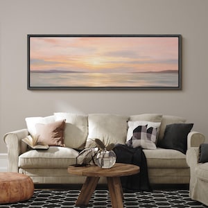 May include: A framed landscape painting depicts a serene sunset over water, with soft pink, orange, and blue hues. The painting hangs above a beige sofa adorned with pillows, a wooden coffee table, and a patterned rug, creating a cozy living room setting.