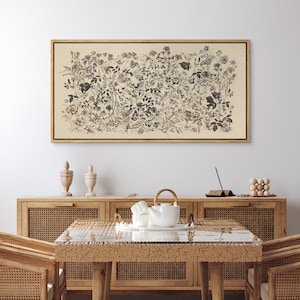 May include: A framed botanical print featuring a detailed black and white illustration of various flowers and foliage. The artwork is set against a cream-colored background and encased in a thin, gold-colored frame. The print is displayed above a wooden cabinet.