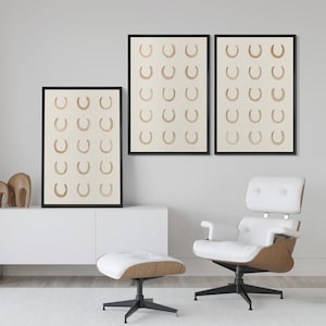 May include: Three framed art prints featuring a repeating pattern of brown horseshoes on a cream background. The prints are displayed in black frames. The room also includes a white chair and ottoman.