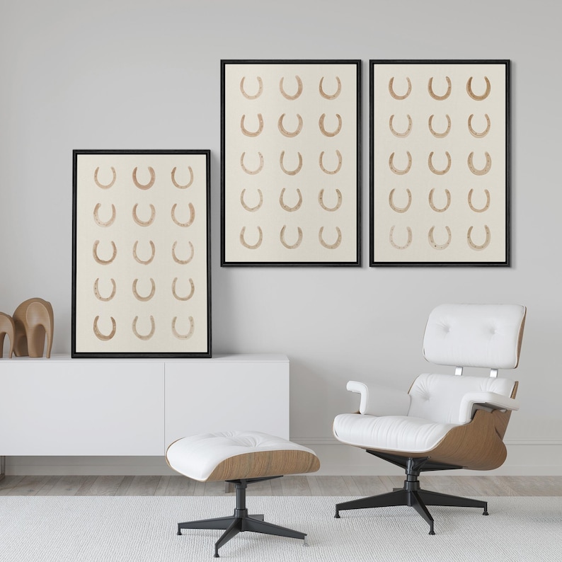 May include: Three framed art prints featuring a repeating pattern of brown horseshoes on a cream background. The prints are displayed in black frames. The room also includes a white chair and ottoman.