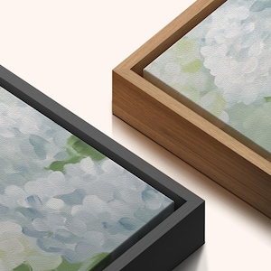 Puede incluir: Dos obras de arte en lienzo con pinturas florales. Una est&aacute; enmarcada en un marco de madera negro, la otra en un marco de madera natural. Las pinturas presentan suaves tonos azules, blancos y verdes, creando una est&eacute;tica serena y art&iacute;stica.